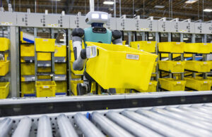 Agility Robotics' Digit humanoid robot carrying a yellow bin in front of a conveyor belt. Ricoh is taking over commercial support.