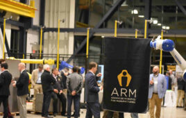 A white and blue robotic arm holding up an ARM Institute flag in front of a crowd of people at the ARM Institute.