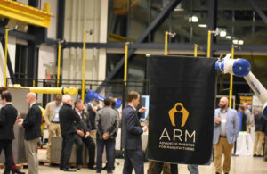 A white and blue robotic arm holding up an ARM Institute flag in front of a crowd of people at the ARM Institute.