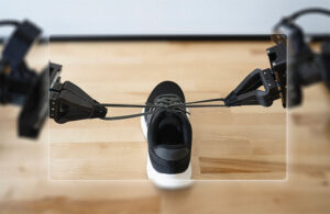 Two black robotic arms finish tying shoelaces into a bow on a black and white pair of shoes.