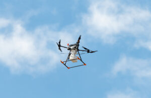 A DroneUp drone flying in the sky with clouds behind it.