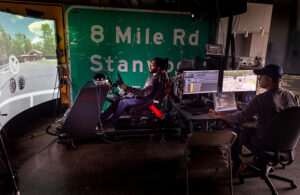 One man sits, reclined as a driver would, with a steering wheel and dashboard set up immediately in front of him, along with a large screen further away that gives a view from the driver's perspective on a remote controlled car. To the right, sits another man at a desk behind a bank of computer screens at Mcity.