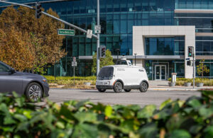 A small, boxy, white Nuro vehicle driving on a road with a glass building behind it.