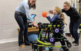 A young girl with long blonde hair using the Trexo Robotics brace to walk and play with a basketball.