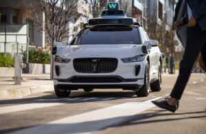 A white Waymo vehicle with the Waymo sensor stack on top. The car is stopped at an intersection with a pedestrian walking across the road in front of it. VERSES AI and Volvo are working to predict such occurrences.