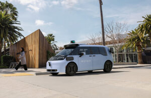 A picture of Waymo's 6th generation vehicle, an white boxy Zeekr vehicle with Waymo's branded sensor stack on top, in front of a white and wood building with palm trees around.