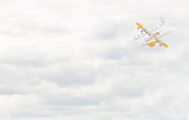 A yellow and white Wing drone in the upper right corner, flying into a cloudy sky.